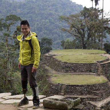 An American Chinese man with a prosthetic leg standing in a epic green landscape and stone built up platforms with paths on Inca Island