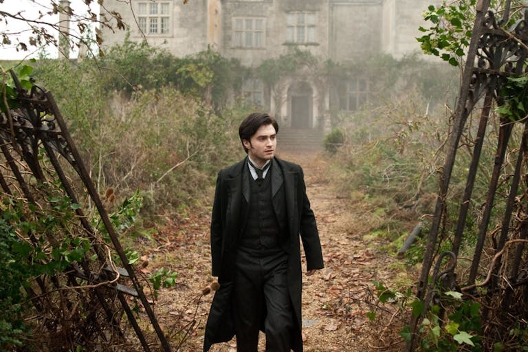 A white man with period era suit and long jacket walks through a rundown and heavily weeded grand estate through broken and rusted metal gates