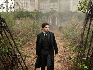 A white man with period era suit and long jacket walks through a rundown and heavily weeded grand estate through broken and rusted metal gates