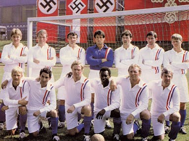 A football team and coach of 13 men in two rows, one standing, one kneeling in front of a football goal and fascist flags hung behind them