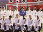 A football team and coach of 13 men in two rows, one standing, one kneeling in front of a football goal and fascist flags hung behind them