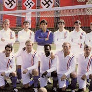A football team and coach of 13 men in two rows, one standing, one kneeling in front of a football goal and fascist flags hung behind them