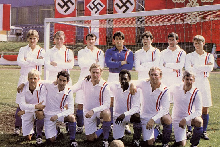 A football team and coach of 13 men in two rows, one standing, one kneeling in front of a football goal and fascist flags hung behind them