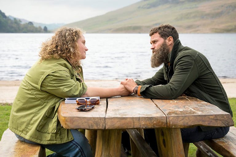 A white woman with curly blonde hair and a white man with dark hair and beard, sit at a picnic bench beside a lake holding hands across the table