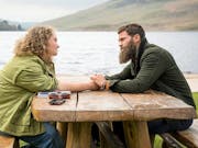 A white woman with curly blonde hair and a white man with dark hair and beard, sit at a picnic bench beside a lake holding hands across the table