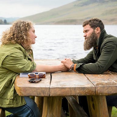 A white woman with curly blonde hair and a white man with dark hair and beard, sit at a picnic bench beside a lake holding hands across the table