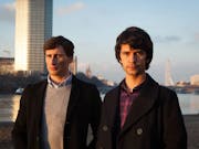Two young man stand outside against a city backdrop
