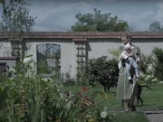A white woman carrying a baby walking in a large, bright enclosed garden