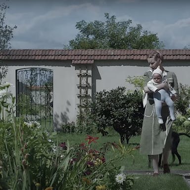 A white woman carrying a baby walking in a large, bright enclosed garden
