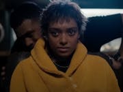 Young woman in a yellow hoodie staring straight ahead with a man in the shadows looking over her shoulder