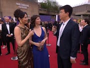Three young people in elegant dresses and a suit stand on a red carpet, the Southbank Centre in the background, with many people in suits around them