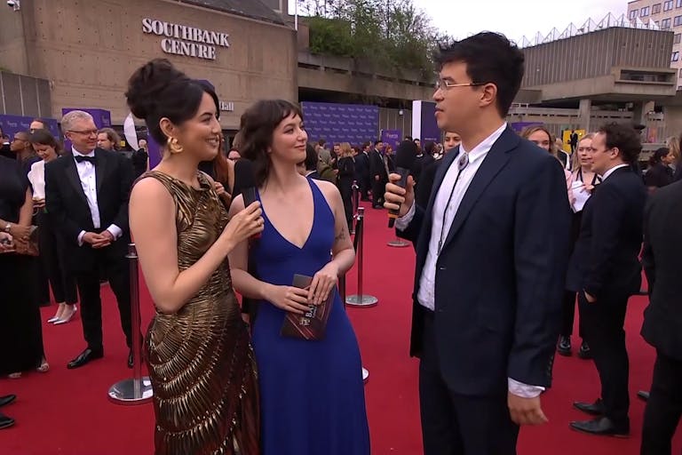 Three young people in elegant dresses and a suit stand on a red carpet, the Southbank Centre in the background, with many people in suits around them 