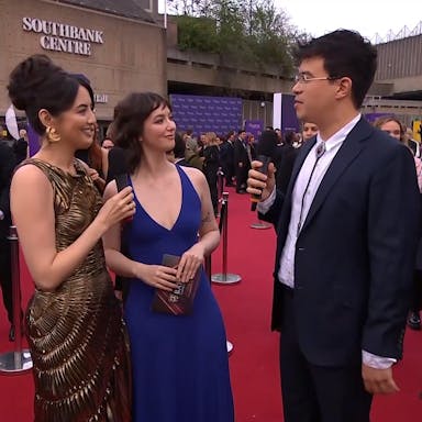 Three young people in elegant dresses and a suit stand on a red carpet, the Southbank Centre in the background, with many people in suits around them 