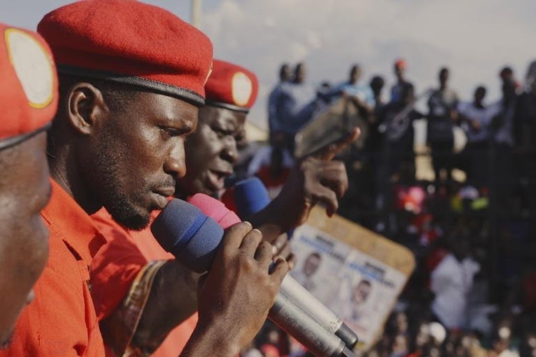 Three Black men in red shirts and red berets speaking into microphones to a crowd
