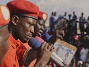 Three Black men in red shirts and red berets speaking into microphones to a crowd