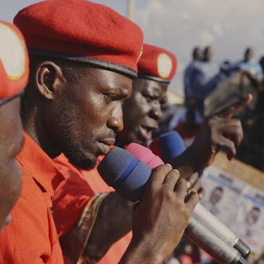 Three Black men in red shirts and red berets speaking into microphones to a crowd