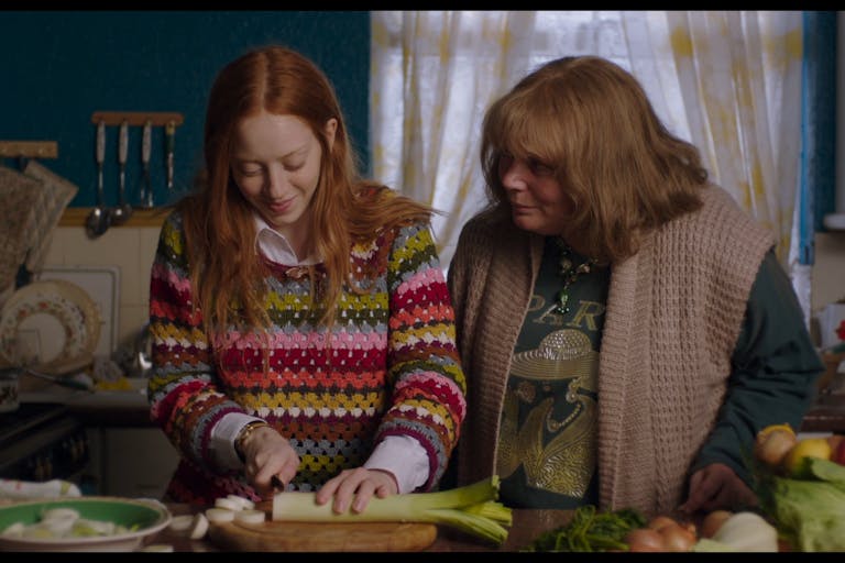 A teenage girl wearing a multi-coloured jumper in a kitchen with her mum chopping a leek