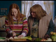 A teenage girl wearing a multi-coloured jumper in a kitchen with her mum chopping a leek