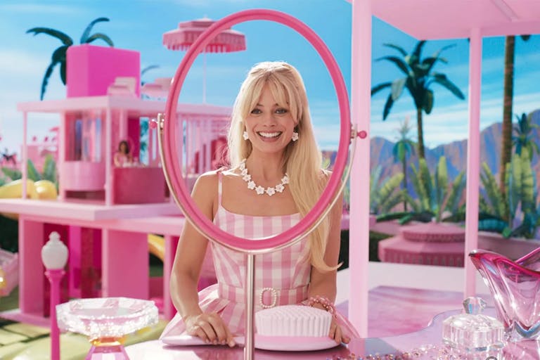 A blonde woman smiling widely, looking through an oval frame with missing mirror in a Barbie pink environment