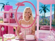 A blonde woman smiling widely, looking through an oval frame with missing mirror in a Barbie pink environment