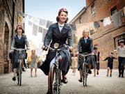 Three young white women in period nurse uniforms riding bikes with young kids playing in the background