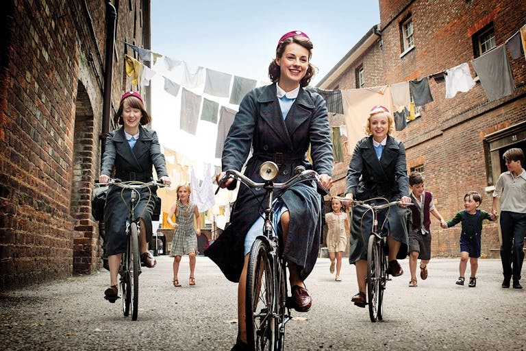 Three young white women in period nurse uniforms riding bikes with young kids playing in the background