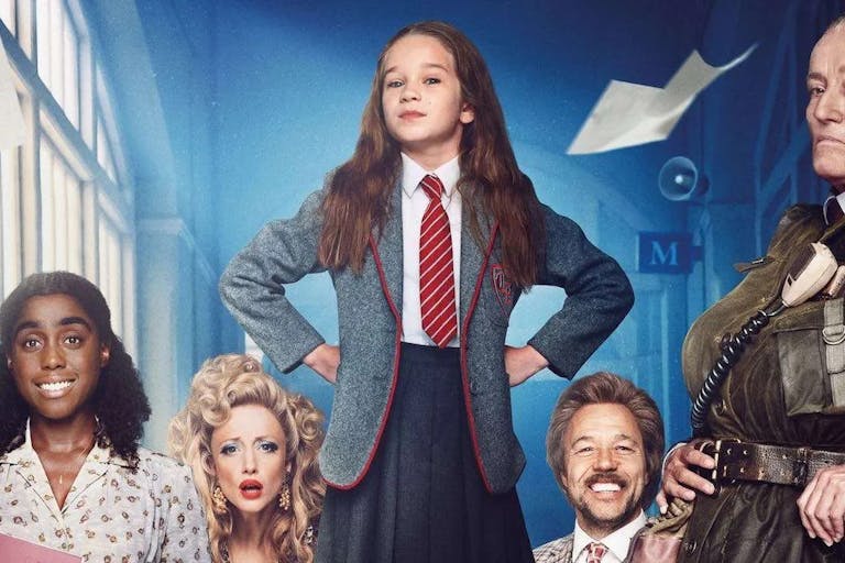 Young girl in school uniform, a red tie and grey blazer, stands with her hands on her hips looking defiantly at the camera, surrounded by other people.