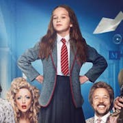 Young girl in school uniform, a red tie and grey blazer, stands with her hands on her hips looking defiantly at the camera, surrounded by other people.