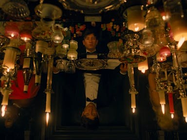 A young white man in a tux sitting at a grand table with many candles covering the table, his image reflected in a disorientating way