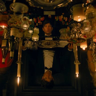 A young white man in a tux sitting at a grand table with many candles covering the table, his image reflected in a disorientating way