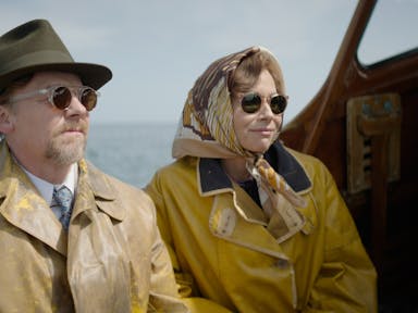 A middle aged man and woman sitting on a small boat wearing mustard yellow rain coats, dark sunglasses, a fedora and silk headscarf 