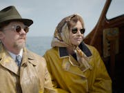 A middle aged man and woman sitting on a small boat wearing mustard yellow rain coats, dark sunglasses, a fedora and silk headscarf