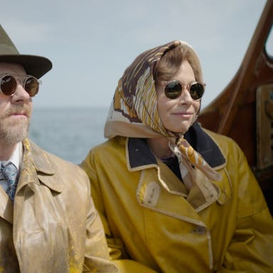 A middle aged man and woman sitting on a small boat wearing mustard yellow rain coats, dark sunglasses, a fedora and silk headscarf 
