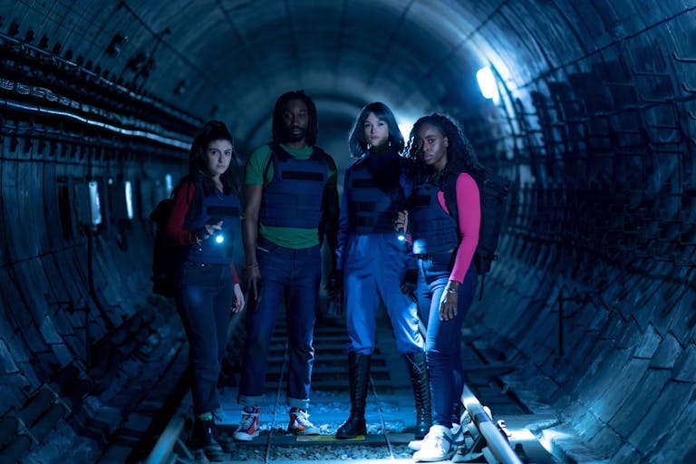 A Black man and woman and two white women stand in a dark underground train tunnel wearing bulletproof vests