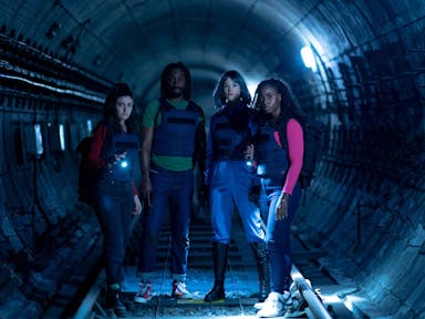 A Black man and woman and two white women stand in a dark underground train tunnel wearing bulletproof vests