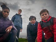 Four teenagers, three boys and a girl, in their raincoats against a grey sky, look worriedly at the camera.