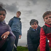 Four teenagers, three boys and a girl, in their raincoats against a grey sky, look worriedly at the camera.