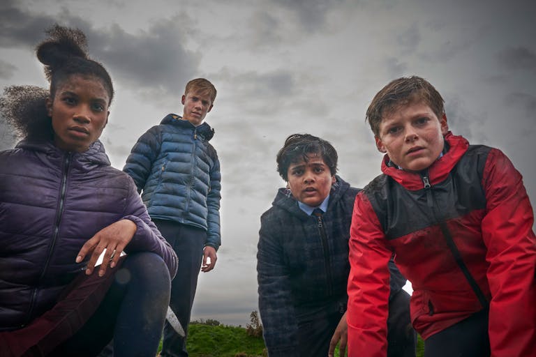 Four teenagers, three boys and a girl, in their raincoats against a grey sky, look worriedly at the camera. 