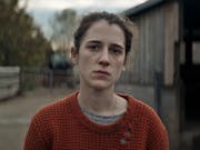 Young woman with dark hair pulled back, wearing a holey orange jumper, standing in a farm