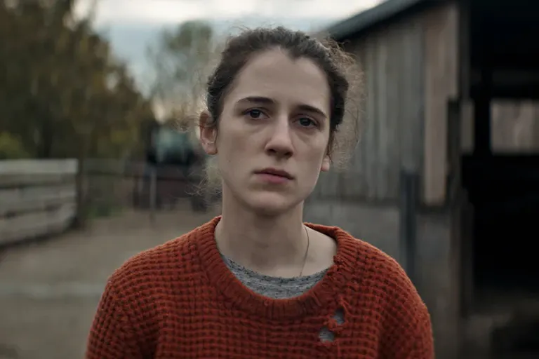Young woman with dark hair pulled back, wearing a holey orange jumper, standing in a farm