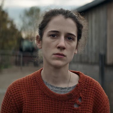 Young woman with dark hair pulled back, wearing a holey orange jumper, standing in a farm 