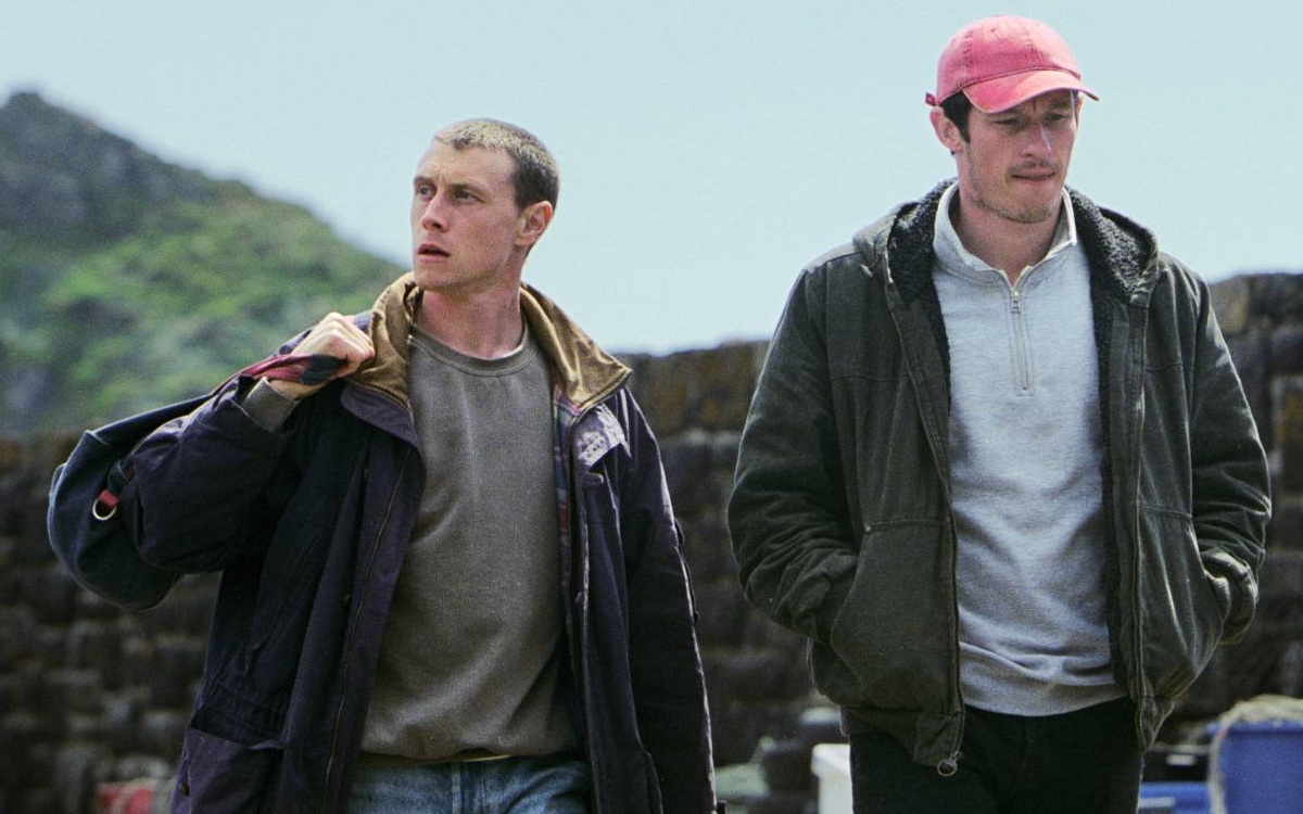 Two early 30's white men walking next to eachother away from hills in the background, both of which wearing a dark jacket. One looking down, the other (holding a bag) looking away