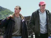 Two early 30's white men walking next to eachother away from hills in the background, both of which wearing a dark jacket. One looking down, the other (holding a bag) looking away