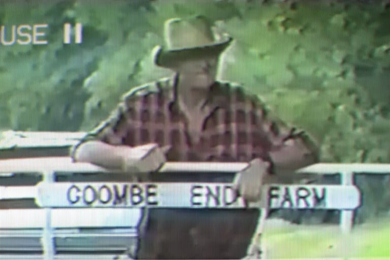 An old farmer in a red plaid shirt and brown brimmed hat, leaning against a fence with with sign reading Coombe End Farm attached