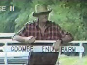 An old farmer in a red plaid shirt and brown brimmed hat, leaning against a fence with with sign reading Coombe End Farm attached