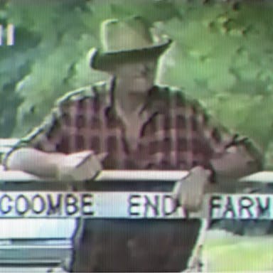 An old farmer in a red plaid shirt and brown brimmed hat, leaning against a fence with with sign reading Coombe End Farm attached