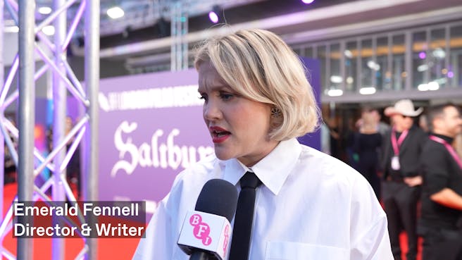 A blonde white woman in white shirt and skinny black tie speaks into a microphone on a red carpet