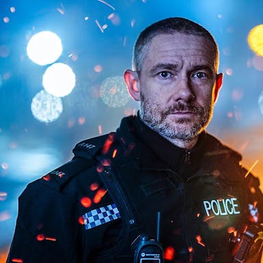 Man in British police uniform looks at the camera against the backdrop of city lights at night. 