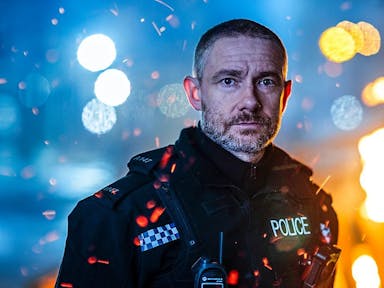 Man in British police uniform looks at the camera against the backdrop of city lights at night. 
