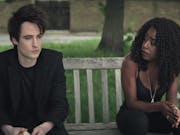 A man and a woman sit on a bench a little distance apart looking sombre